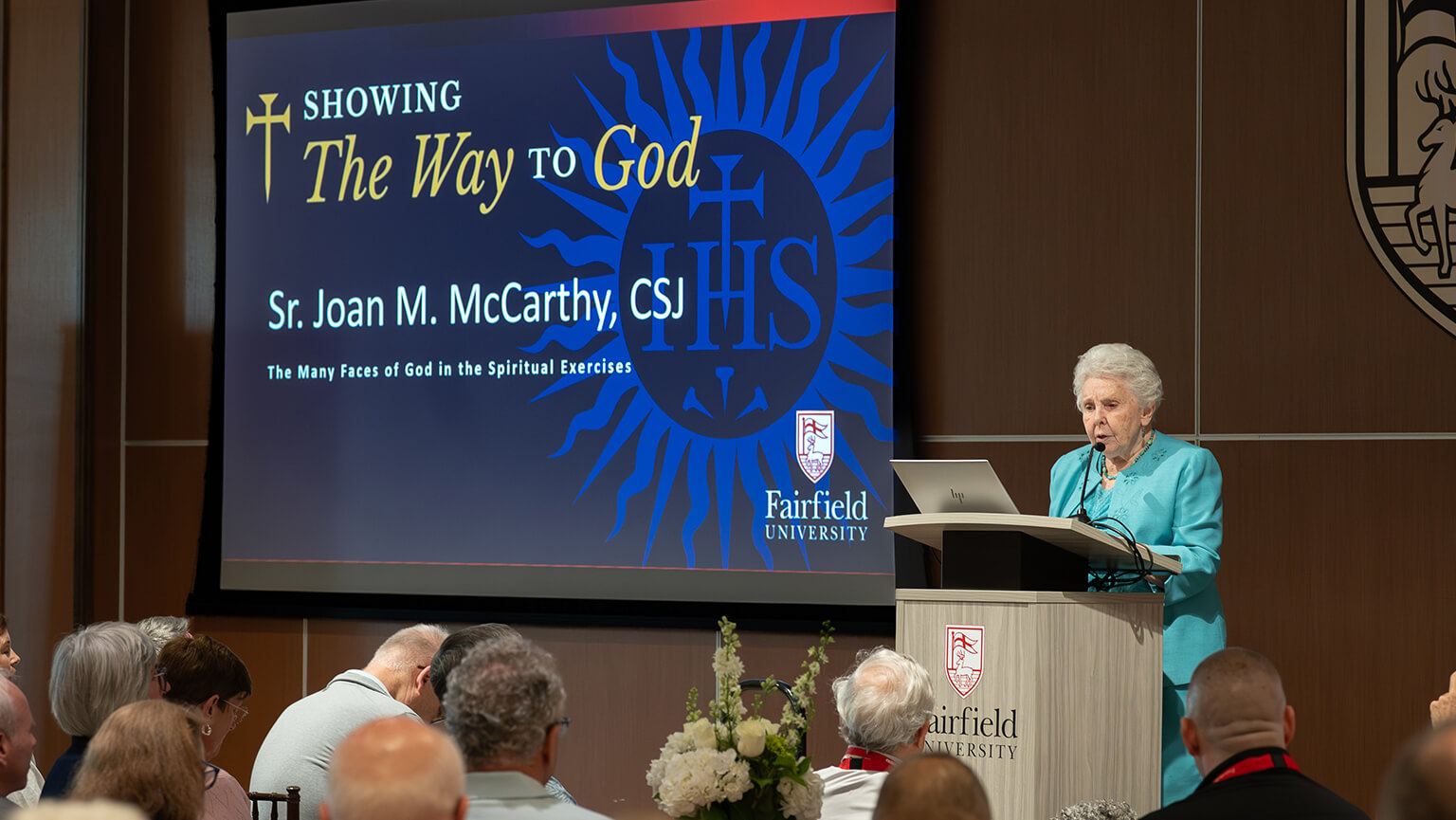 Sr. Joan McCarthy, CSJ presents a lecture titled “The Way to God,” featuring blue religious imagery, while seated attendees listen in a warm auditorium.
