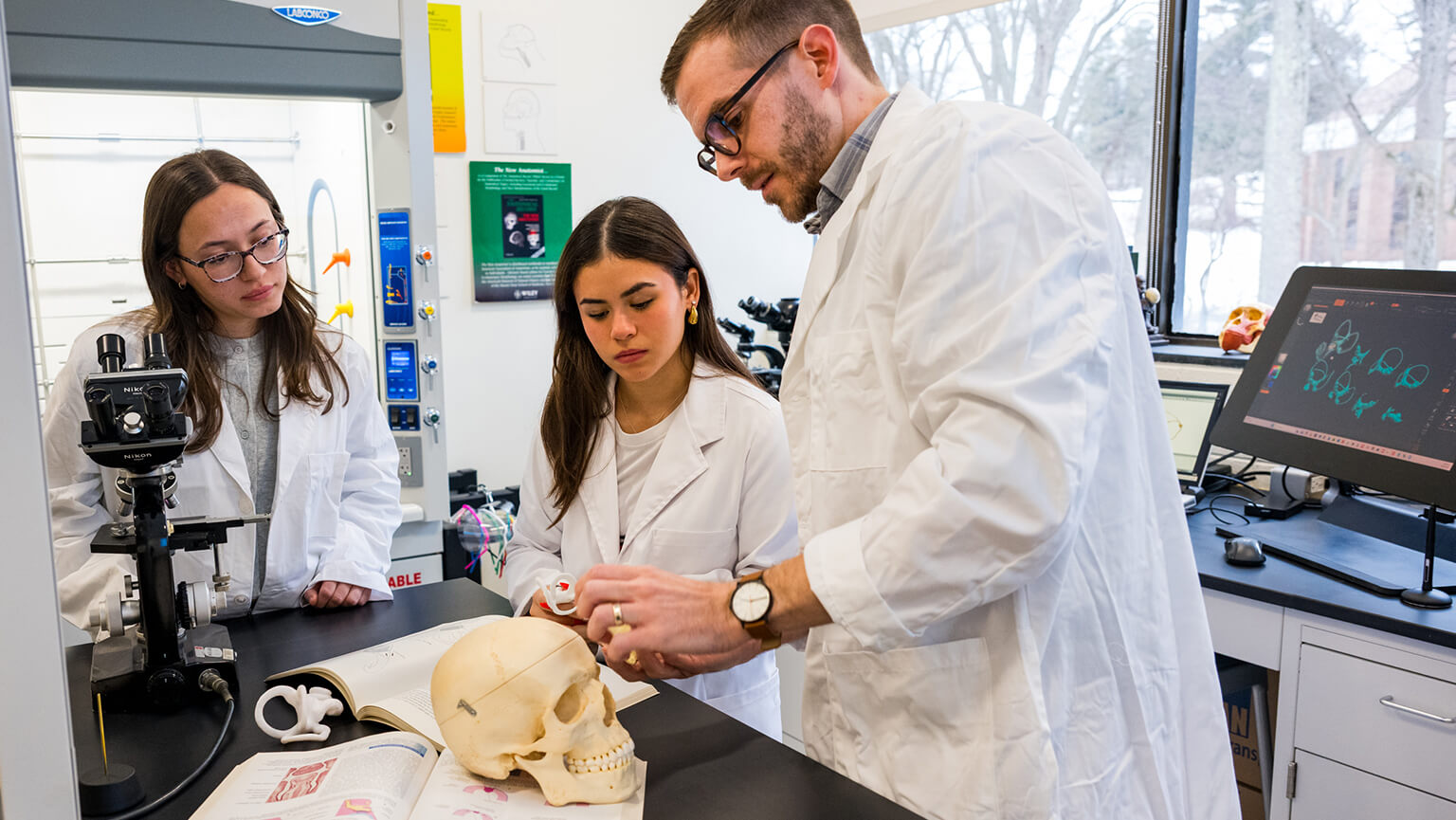 Two students and a bearded instructor examine a skull in a teaching lab, with microscopes and computer screens nearby; the scene feels focused and educational.