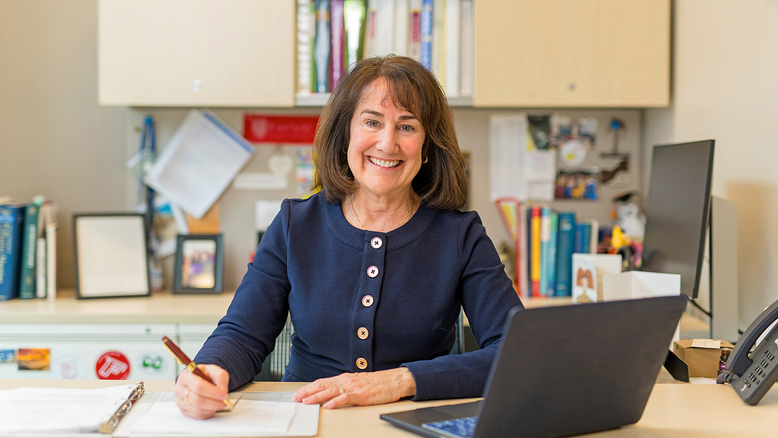 Professor Eileen O’Shea in a navy blouse, smiling warmly, is seated at a desk with a pen and paper. A laptop is open nearby. Books and files are visible in the background, suggesting a professional setting.
