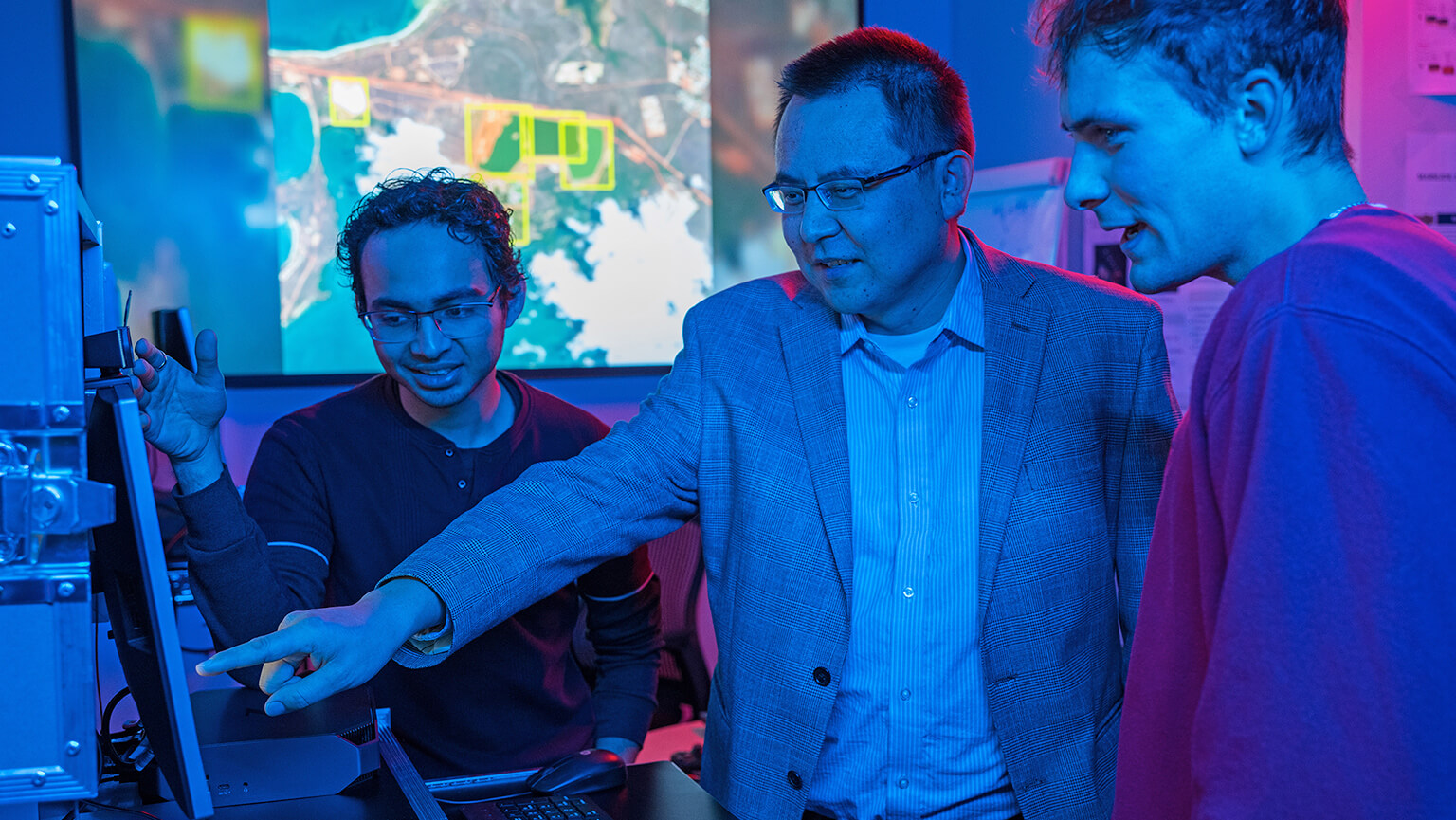 Three people collaborate in a dimly lit room with blue lighting. One points at a computer screen. A map is displayed in the background, signifying tech work.