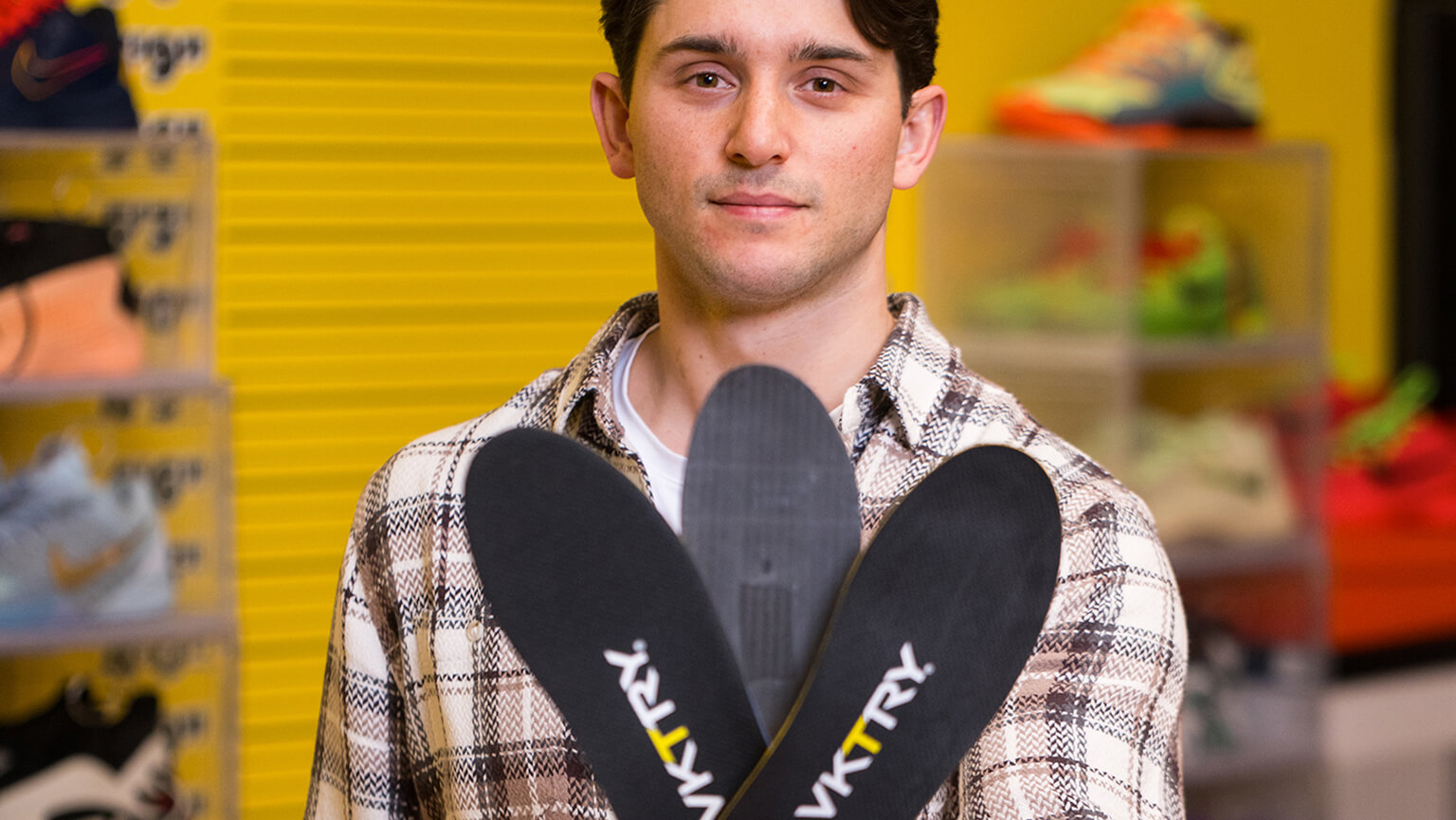 Drew Aricuolo ’18 in a plaid shirt holds three black insoles labeled "VKTRY" in a sports store. The background features colorful athletic shoes, creating an energetic tone.
