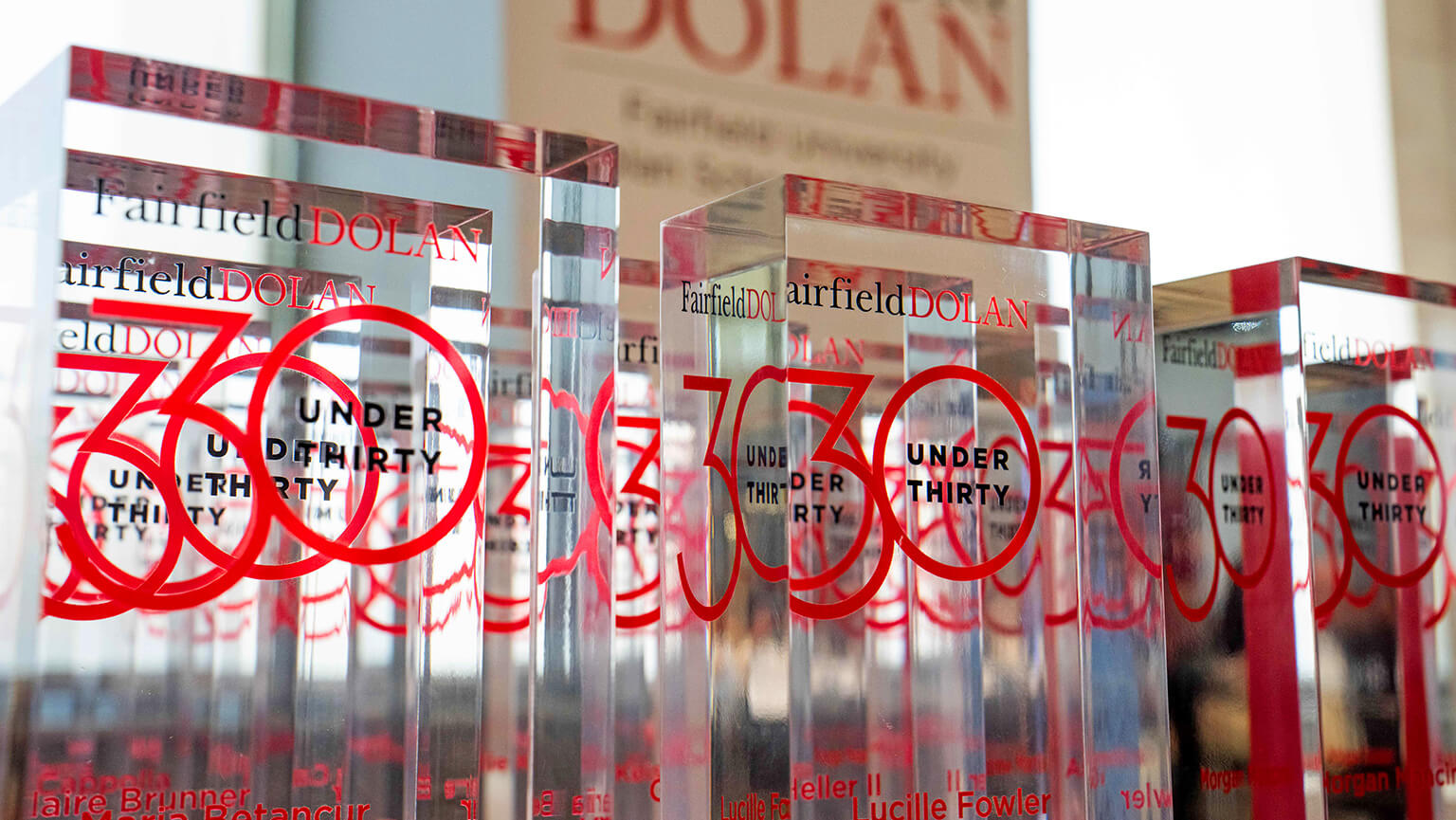 A row of clear glass awards with red "30 Under Thirty" text showcasing Fairfield Dolan branding. The awards are sleek and modern, conveying prestige.