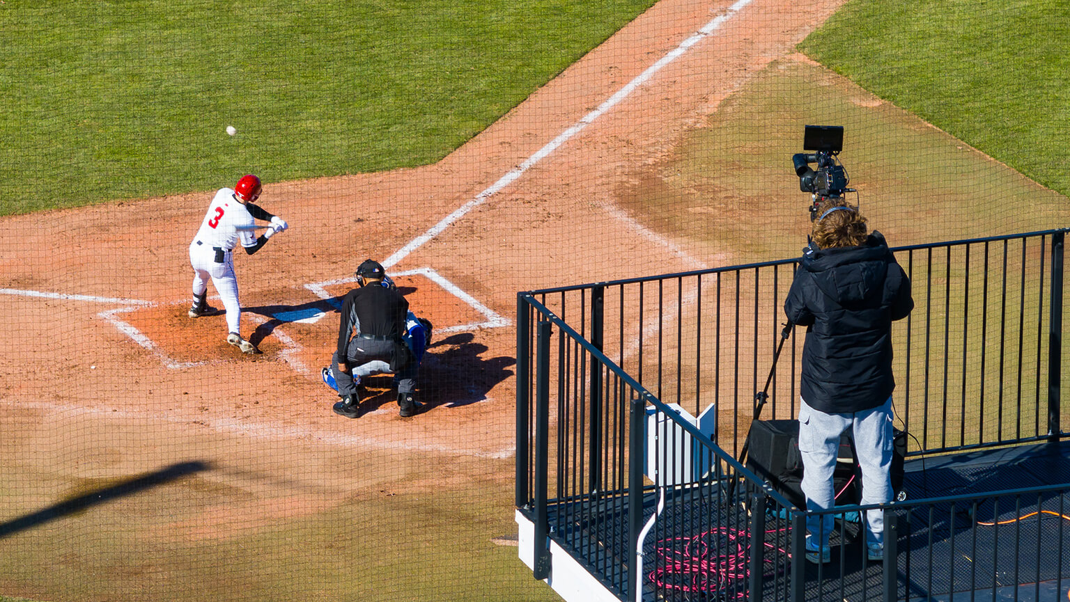 Aerial view of a baseball game with a batter in a red helmet ready to hit. A catcher and umpire are crouched behind. A camera operator films nearby.