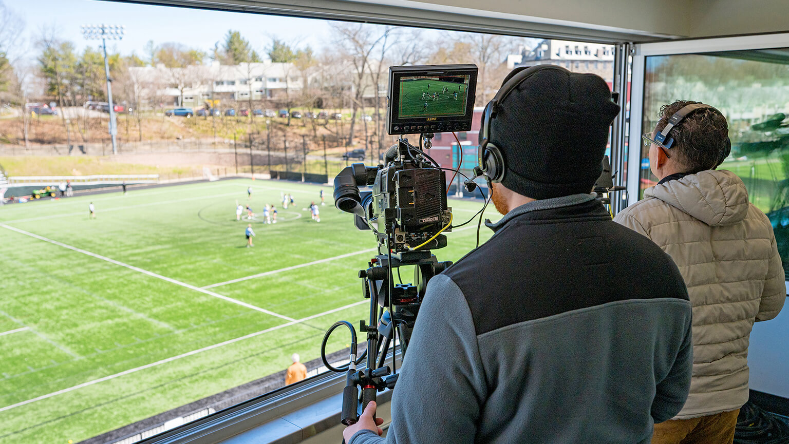 Two cameramen film a soccer game from a press box overlooking a green field. One operates a camera with a monitor, capturing the action below.