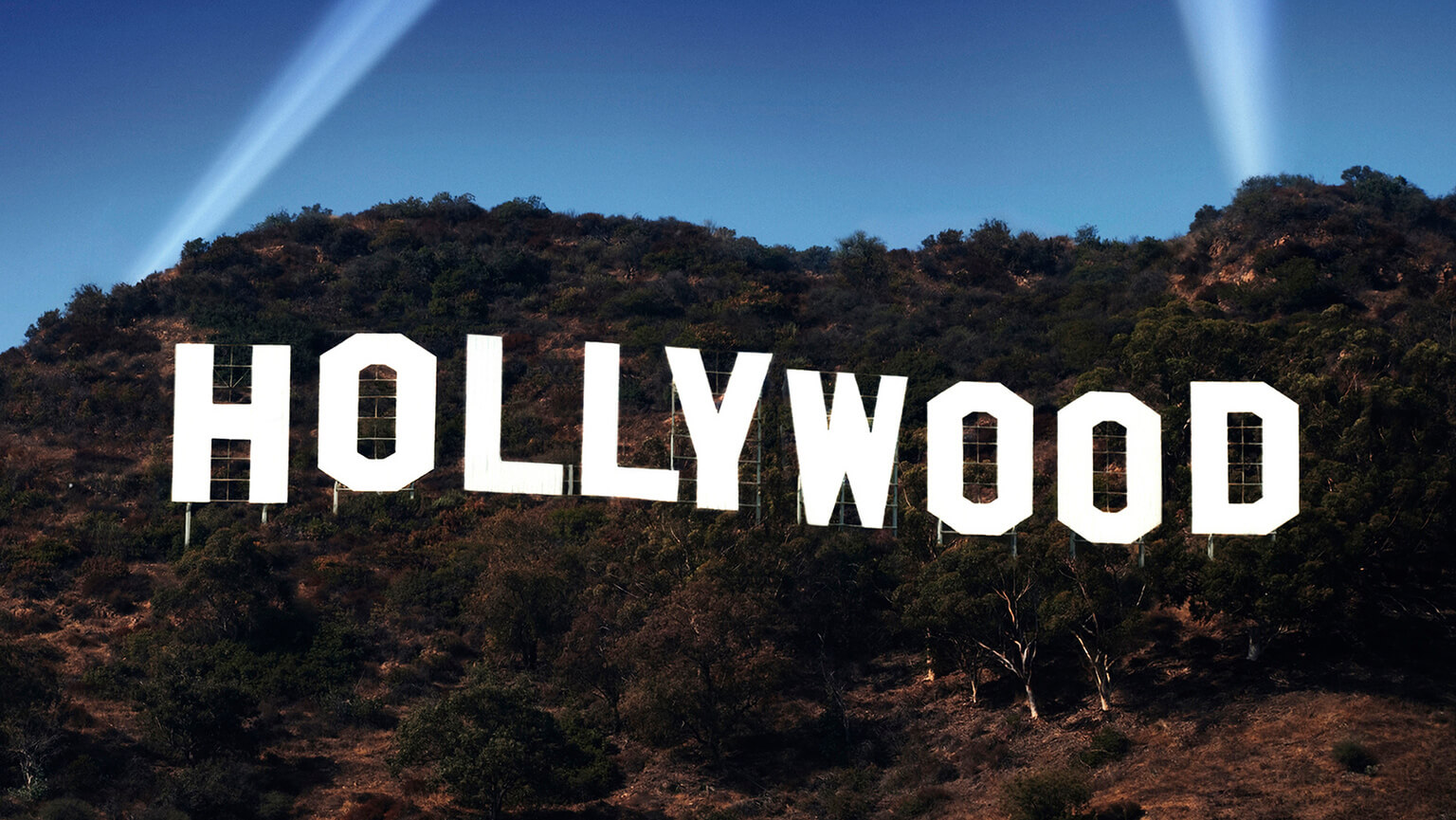 Famous white "Hollywood" sign on a brown hillside with green trees, under a clear blue sky, illuminated by beams of light. Iconic, cinematic atmosphere.