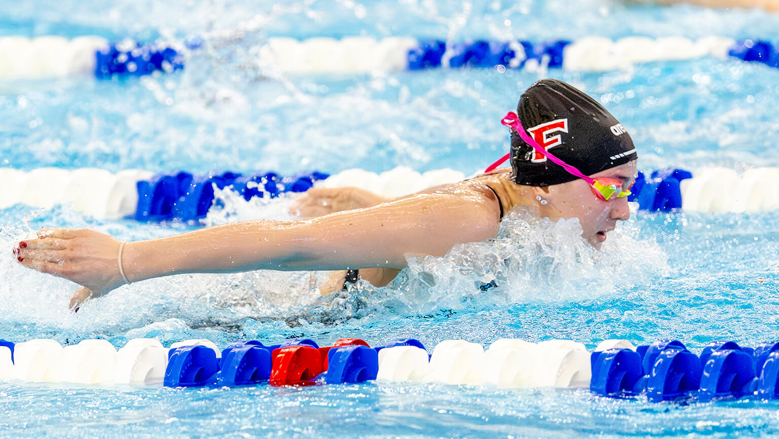 Riley Edge ’29 in a black cap and goggles competes in a butterfly stroke race, surrounded by blue water and colorful lane dividers, conveying focus and determination.