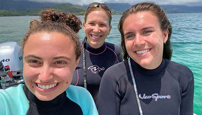 Three women in wetsuits smiling happily at the camera, showcasing their excitement for water activities.