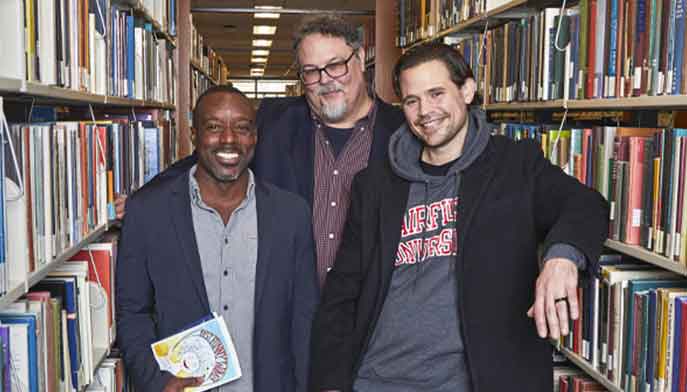 Three men standing in a library, positioned in front of tall bookshelves filled with various books.