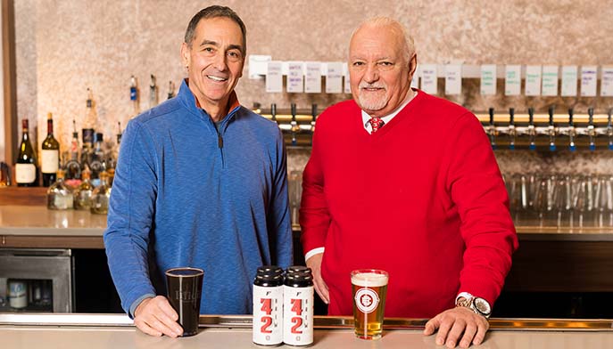 Image of Two friendly men at a bar, standing behind a counter filled with various beers and drinks.