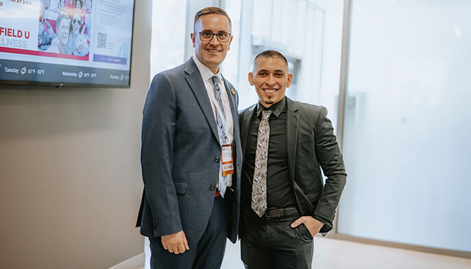 Two men in suits standing side by side, both smiling and looking towards the camera. 