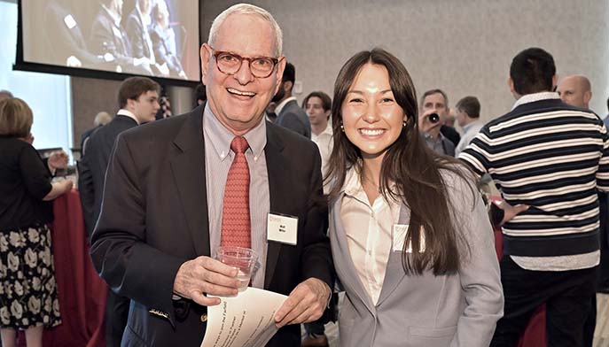 Image of two people standing side by side, smiling at an event, with a crowd and decorations in the background.