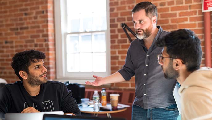Image of three men sit around a table, each focused on their laptops, engaged in a collaborative discussion.