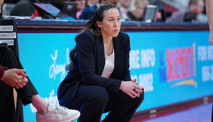 Head coach of the Fairfield University woman's basketball team dressed in a suit sits on the sidelines, attentively watching the proceedings unfold.