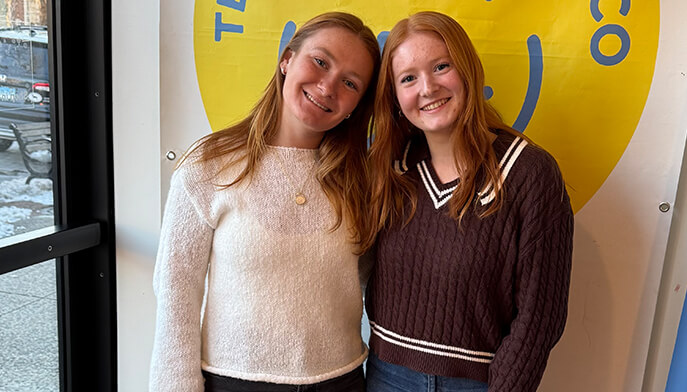 Two young women stand side by side in front of a bright yellow sign, smiling and looking at the camera.