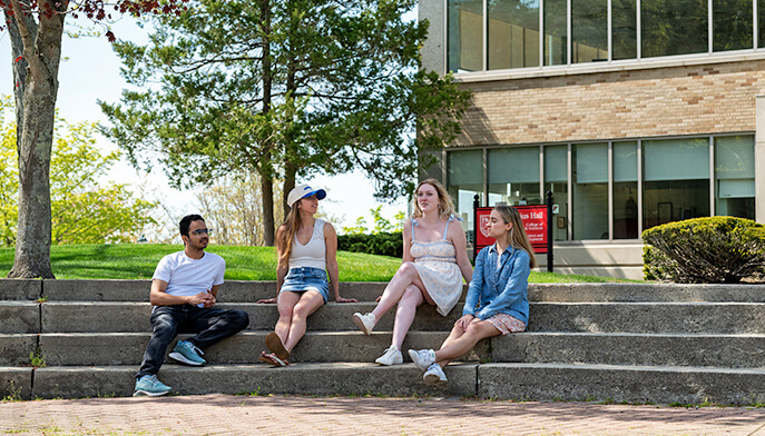Four students relax on the steps of a building, interacting with each other in a friendly and informal atmosphere.  