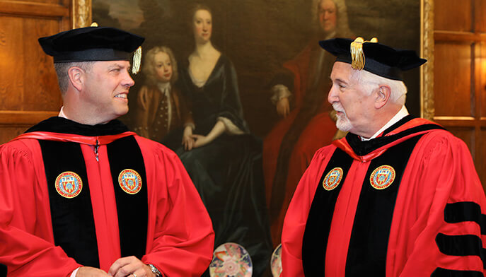 Two men wearing red robes and black hats stand side by side, showcasing a traditional or ceremonial attire.