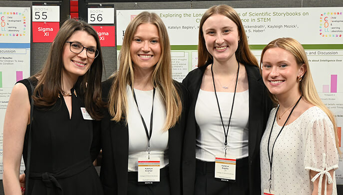 Four women stand together in front of a colorful poster, smiling and engaging with each other. 