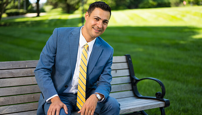 Dario Shore EdD’25 smiling in a blue suit and yellow striped tie sits on a bench, surrounded by green lawn and trees in bright sunlight.