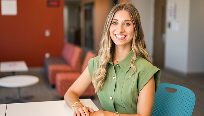 Smiling with long blonde hair, Nicole Dana ’22, MPH’25 sits at a table in a modern room setting, wearing a green blouse beside a blue chair; warm, welcoming portrait.