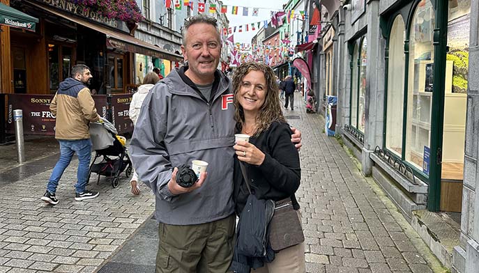Donald and Karen Wilson P’26 A stand arm in arm on a lively cobbled shopping street, holding a camera and takeaway cups beneath colorful flags.