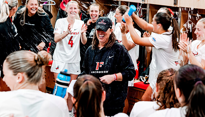 Joyful women’s lacrosse team celebrates in a locker room, spraying water around a laughing coach in a cap and black jacket.