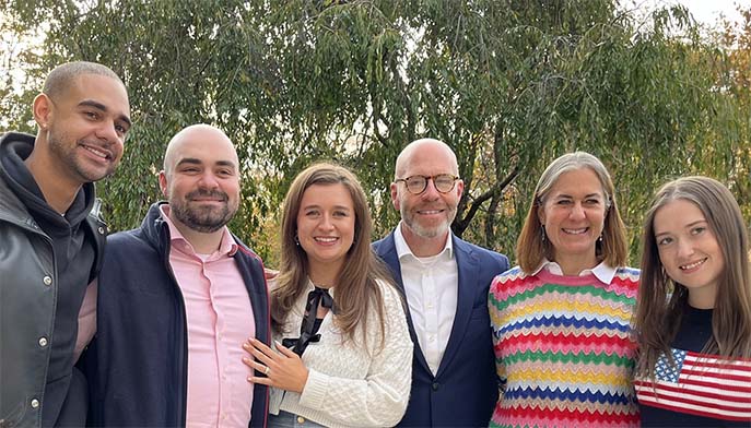 Six smiling adults pose together outdoors beneath leafy trees, dressed in casual and smart clothing, including a bright striped sweater and flag-print top; warm, friendly group portrait.