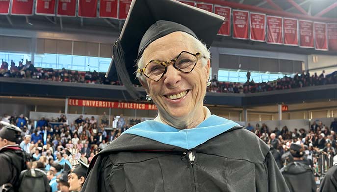 Smiling Ilene Bosch MA’89 in glasses and a black graduation cap and gown with a blue hood stands before a crowd at a university ceremony.