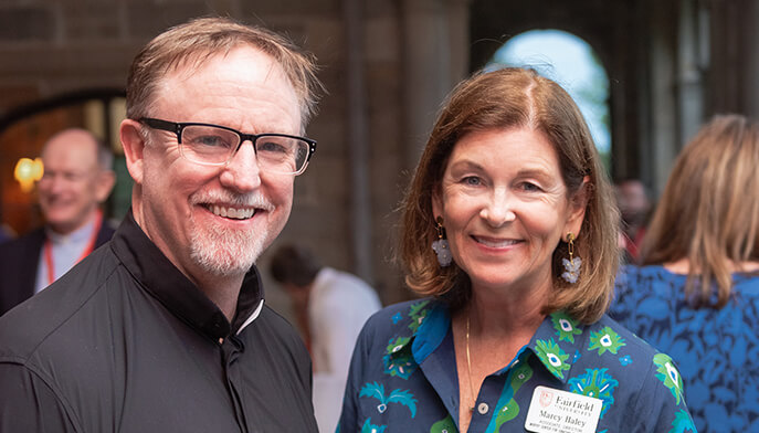 Rev. Denis Donoghue, S.J., in glasses and Marcy Haley in a blue patterned outfit pose together at a crowded indoor event, conveying warmth and camaraderie.
