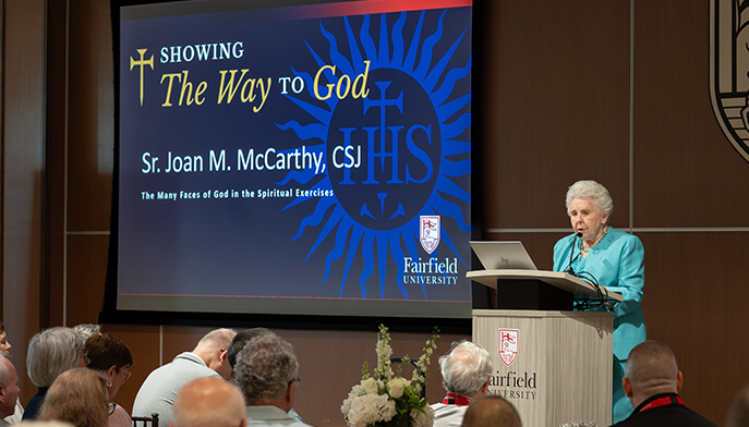 Sr. Joan McCarthy, CSJ presents a lecture titled “The Way to God,” featuring blue religious imagery, while seated attendees listen in a warm auditorium.