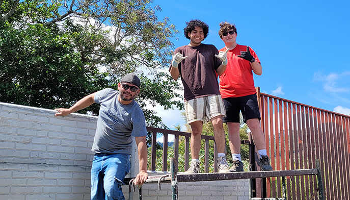 Three students pose on a raised platform by a railing, smiling in casual streetwear under a bright blue sky; the scene feels upbeat and relaxed.
