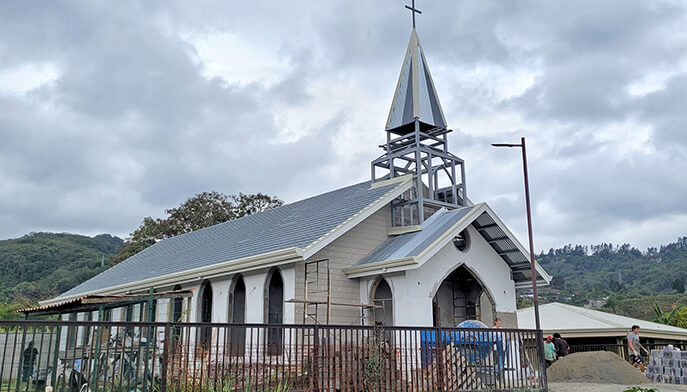 Gray-blue church with a tall steeple and cross stands behind a metal fence, under an overcast sky; the quiet rural scene feels calm and modest.