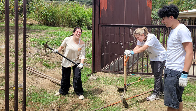 Three students use long-handled tools to clear dirt beside a dark red barn, working together outdoors in a calm, purposeful scene.