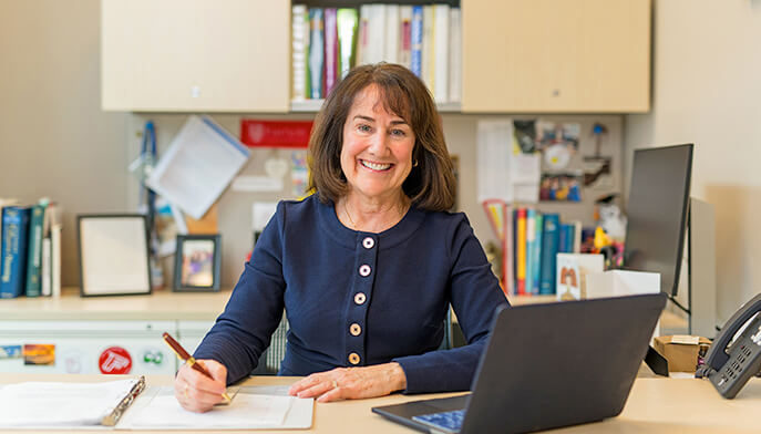 Professor Eileen O’Shea in a navy blouse, smiling warmly, is seated at a desk with a pen and paper. A laptop is open nearby. Books and files are visible in the background, suggesting a professional setting.