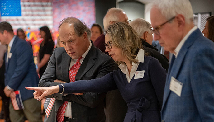 Provost Christine Siegel, PhD points ahead while talking to President Mark R.Nemec, PhD in a crowded event. They appear engaged and serious. Attendees in formal attire are visible in the background.