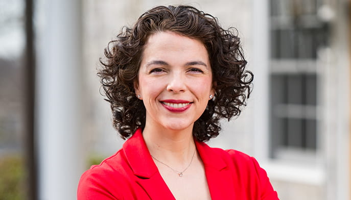 Catherine (Auriana) Cadigan ’04, MBA’10 with curly hair smiling warmly, wearing a red blazer, stands confidently outdoors. Blurred background suggests a professional setting.