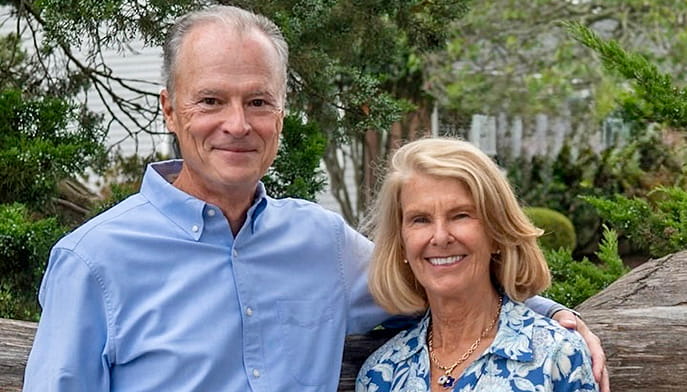 Mike ’75 and Nancy Robinson stand in front of trees with a home in the distance.