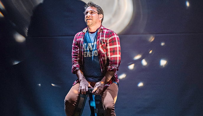Jared Mezzocchi in glasses and a red plaid shirt sits on a stool under bright stage lights against a blurred dark background, conveying a contemplative mood.