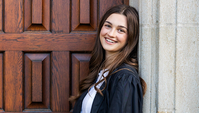 Whitney McVeagh ’25 in a graduation gown happily smiles, leaning against a wooden door. Her relaxed pose and expression convey joy and achievement.