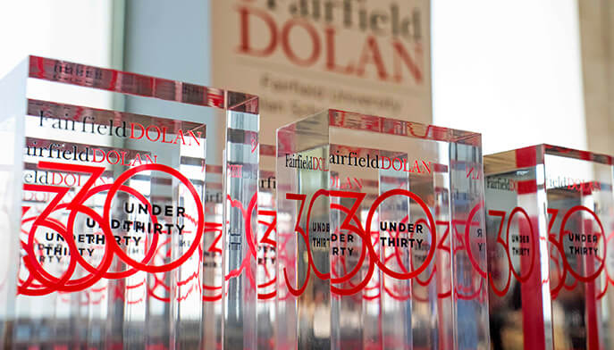 A row of clear glass awards with red "30 Under Thirty" text showcasing Fairfield Dolan branding. The awards are sleek and modern, conveying prestige.