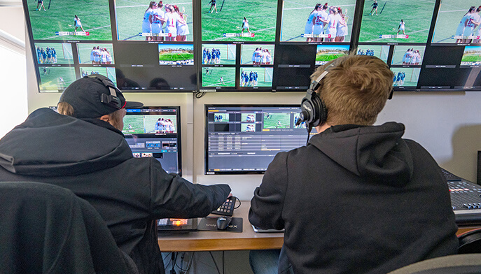 Two people in a control room manage a live sports broadcast. Multiple screens display a soccer match. The atmosphere is focused and professional.