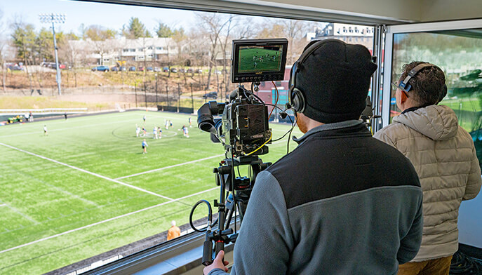 Two cameramen film a soccer game from a press box overlooking a green field. One operates a camera with a monitor, capturing the action below.