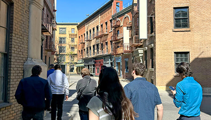 Students walk along a narrow, sunny alley lined with brick buildings and fire escapes. The setting has an urban feel with a hint of exploration.