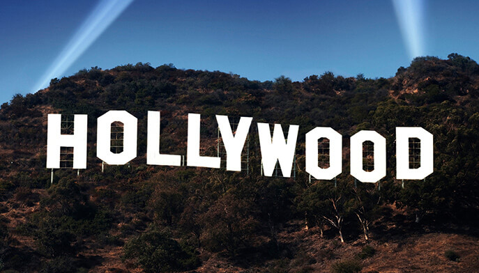 Famous white "Hollywood" sign on a brown hillside with green trees, under a clear blue sky, illuminated by beams of light. Iconic, cinematic atmosphere.