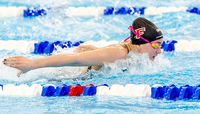 Riley Edge ’29 in a black cap and goggles competes in a butterfly stroke race, surrounded by blue water and colorful lane dividers, conveying focus and determination.