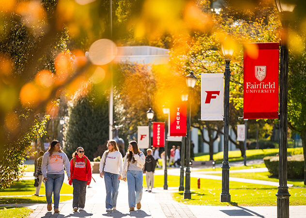 Students walk along sunny Fairfield University campus beneath red banners as golden autumn leaves glow around them, creating a warm, welcoming scene.