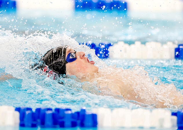 A swimmer, in a black swim cap and blue goggles executes a backstroke with water splashing around. The pool's blue and white lane dividers add contrast.