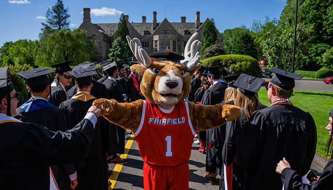 Lucas the Fairfield University mascot wearing a red and black costume and a graduation hat fist bumps students.