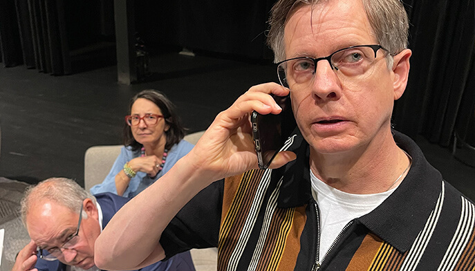 A man seated in a chair, with another man standing next to him engaged in a conversation on his cell phone.
