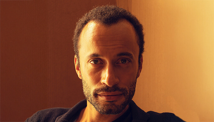 A bearded man dressed in a black shirt, standing against a plain background with a thoughtful expression.