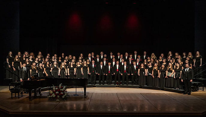 The Glee Club men in tuxedos and women in black dresses stands on tiered risers on the Dr. Carole Ann Maxwell Stage. A grand piano with floral arrangement is in the foreground.